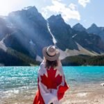 A person in a hat wrapped in a Canadian flag overlooks Moraine Lake and mountains.