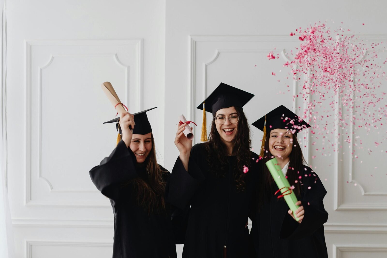 Three young female graduates celebrating indoors with diplomas and confetti.